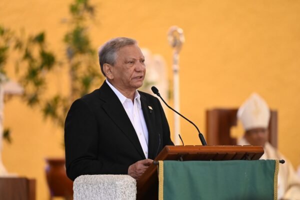 Un hombre de cabello canoso, con saco negro y camisa blanca, habla en un atril de una iglesia mientras un obispo con mitra lo escucha al fondo