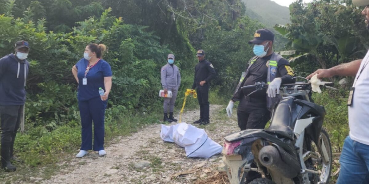 Agentes y personal de salud con mascarilla junto a un cuerpo cubierto con una sábana blanca en un camino de tierra rodeado de vegetación; en primer plano, una motocicleta