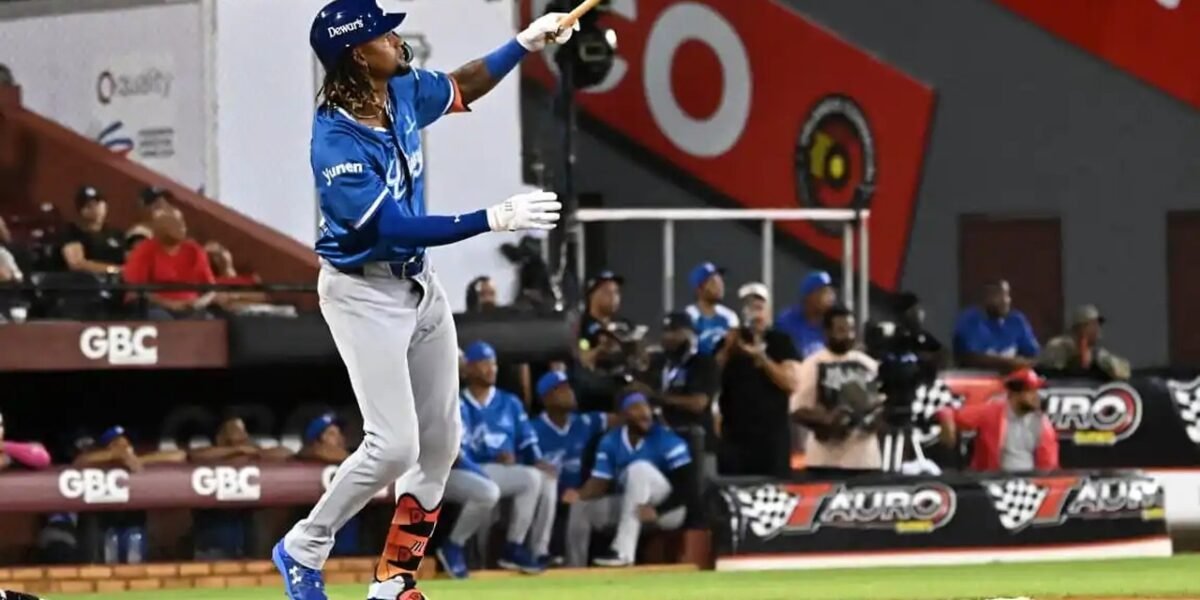 Un bateador de los Tigres del Licey, con uniforme azul y pantalón gris, sigue con la mirada la pelota tras su swing, con compañeros en el dugout al fondo