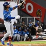 Un bateador de los Tigres del Licey, con uniforme azul y pantalón gris, sigue con la mirada la pelota tras su swing, con compañeros en el dugout al fondo