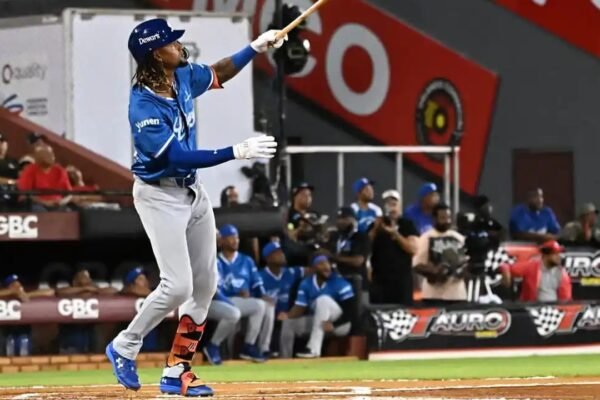 Un bateador de los Tigres del Licey, con uniforme azul y pantalón gris, sigue con la mirada la pelota tras su swing, con compañeros en el dugout al fondo