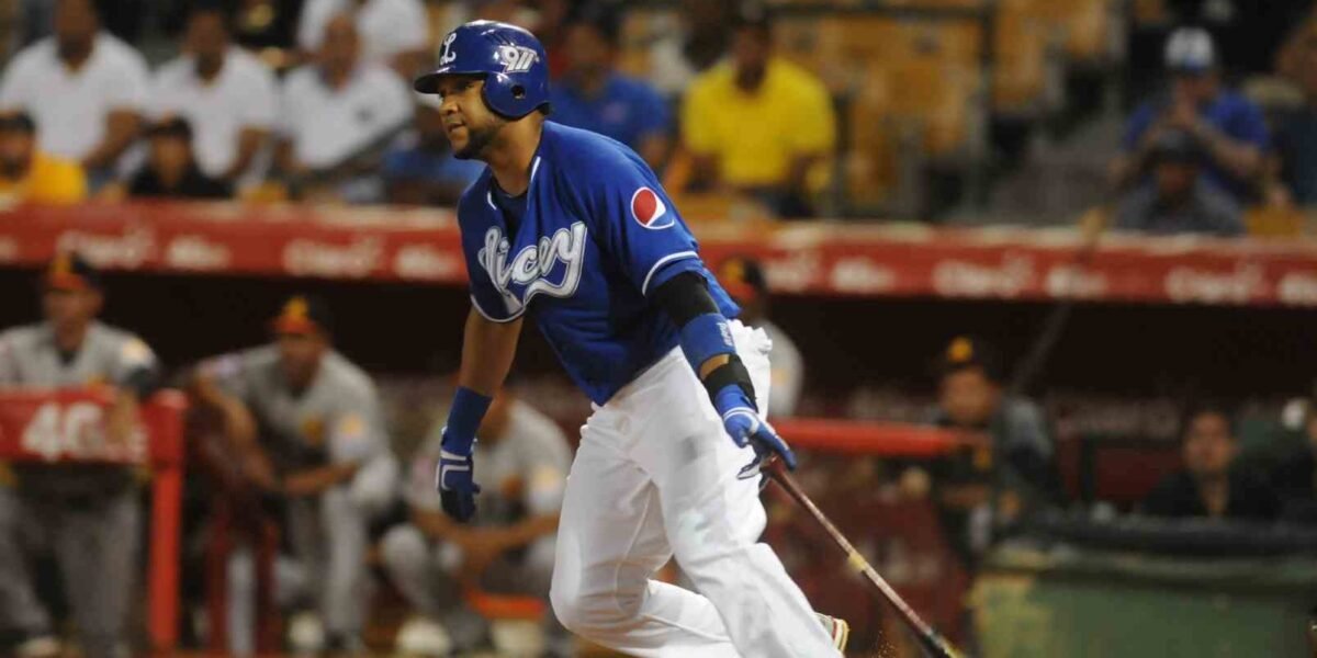Un bateador de los Tigres del Licey, con casco y camiseta azules, suelta el bate y arranca a correr; al fondo, el dugout rival y el público en las gradas