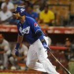 Un bateador de los Tigres del Licey, con casco y camiseta azules, suelta el bate y arranca a correr; al fondo, el dugout rival y el público en las gradas