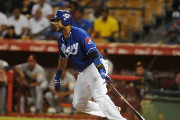 Un bateador de los Tigres del Licey, con casco y camiseta azules, suelta el bate y arranca a correr; al fondo, el dugout rival y el público en las gradas