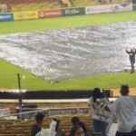 Las lluvias obligan a Lidom a posponer los tres juegos del martes Terreno de un estadio de béisbol cubierto con una lona por la lluvia; dos empleados caminan sobre ella, uno con paraguas rojo, mientras aficionados esperan en las gradas.