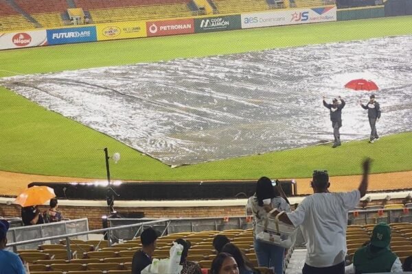 Terreno de un estadio de béisbol cubierto con una lona por la lluvia; dos empleados caminan sobre ella, uno con paraguas rojo, mientras aficionados esperan en las gradas.