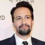Lin-Manuel Miranda, de traje oscuro y barba, posa ante un panel blanco con letras de un acto público
