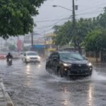 Vehículos y un motorista circulan por una avenida mojada bajo un aguacero junto a un letrero de La Vega; un peatón camina con paraguas por la acera