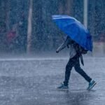 Minerd reanuda este jueves las clases tras levantarse las alertas amarillas Una persona con mochila camina bajo un aguacero cubierta con un paraguas azul sobre una calle mojada.
