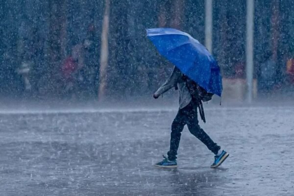 Una persona con mochila camina bajo un aguacero cubierta con un paraguas azul sobre una calle mojada.