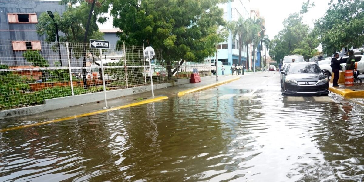 Una calle cubierta de agua junto a una verja metálica y un letrero de una vía; un automóvil negro detenido y varias personas en la acera al fondo