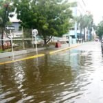 Las lluvias dejan 2,675 afectados y 51 comunidades incomunicadas Una calle cubierta de agua junto a una verja metálica y un letrero de una vía; un automóvil negro detenido y varias personas en la acera al fondo