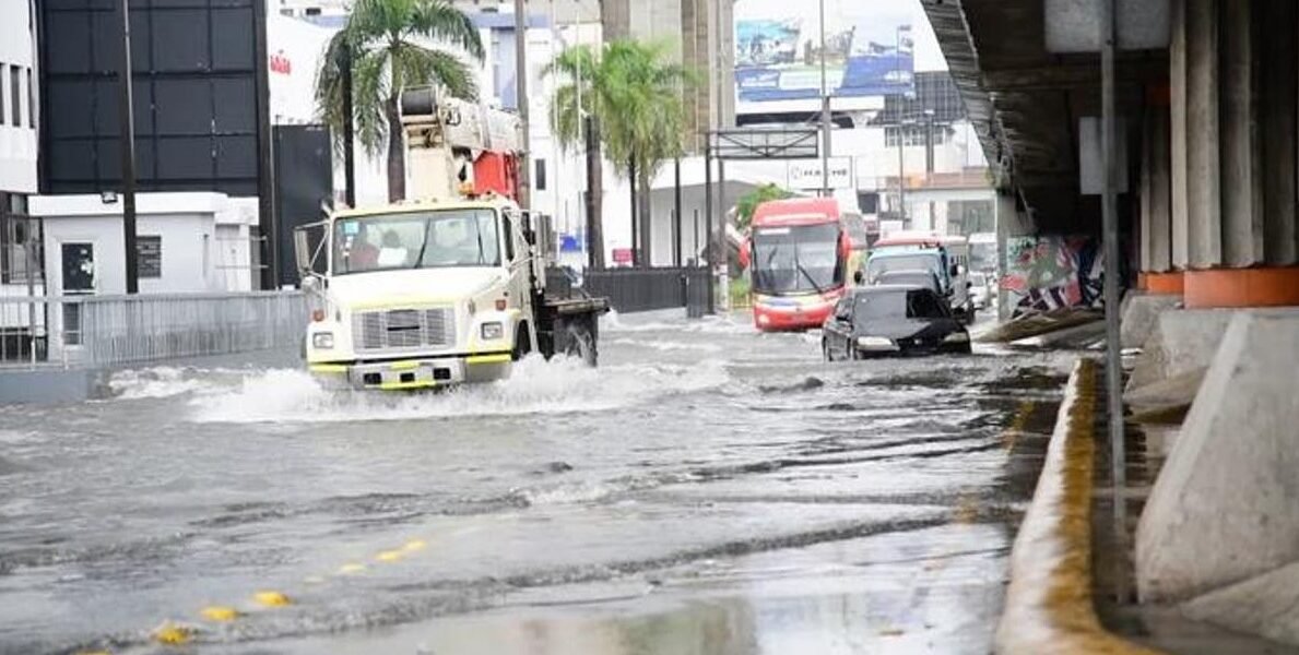 Un camión con grúa, un autobús y varios automóviles circulan por una avenida anegada junto a un paso elevado, bajo un cielo nublado