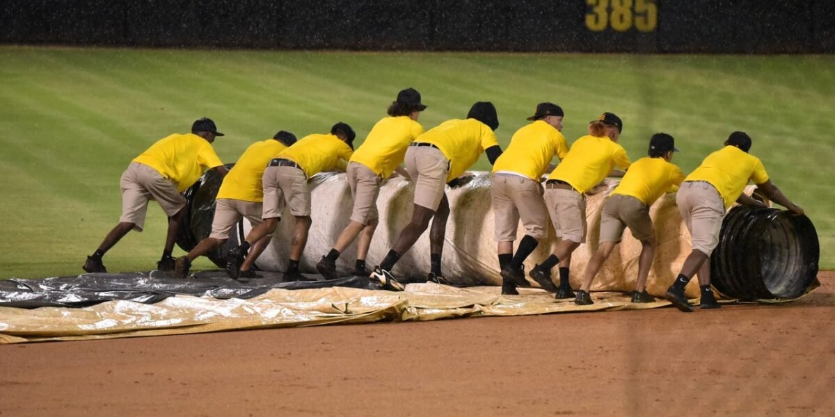 Empleados de mantenimiento con camisetas amarillas despliegan una lona sobre el cuadro de un estadio de béisbol bajo la lluvia