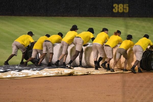 Empleados de mantenimiento con camisetas amarillas despliegan una lona sobre el cuadro de un estadio de béisbol bajo la lluvia