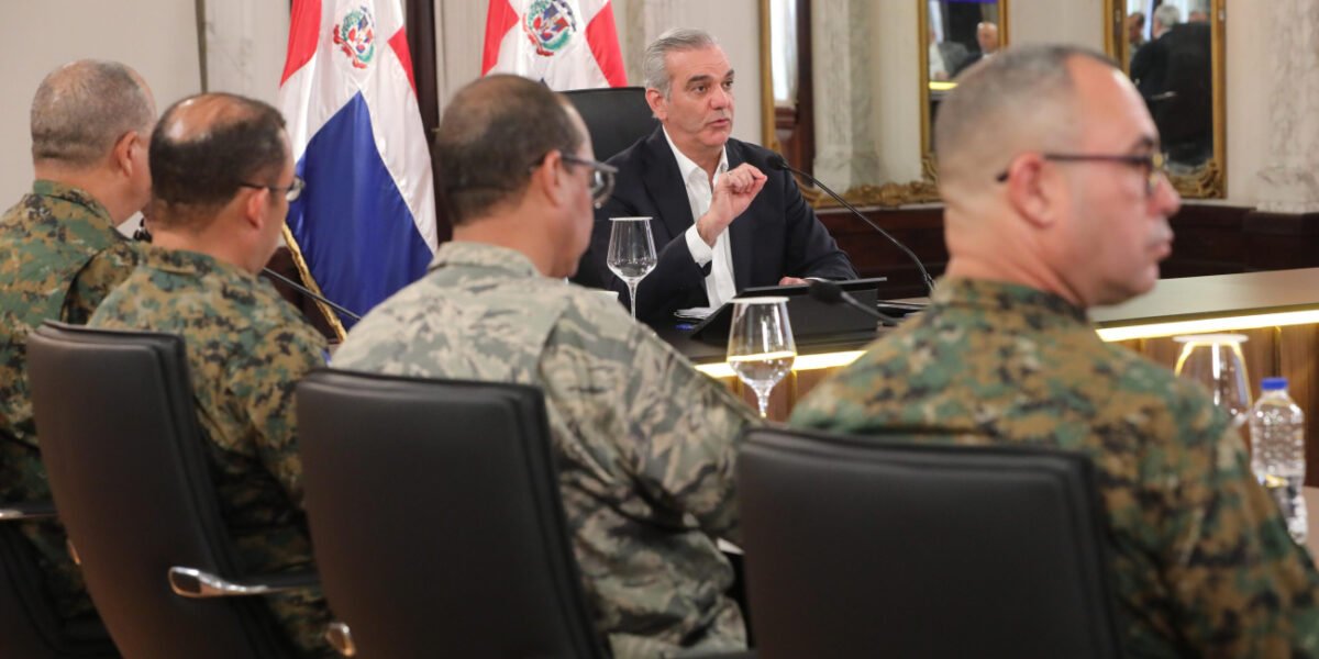El presidente Luis Abinader habla en una mesa frente a varios militares uniformados, con banderas dominicanas al fondo.