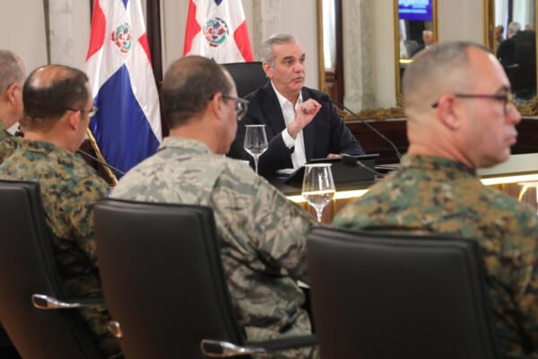 El presidente Luis Abinader habla en una mesa frente a varios militares uniformados, con banderas dominicanas al fondo.