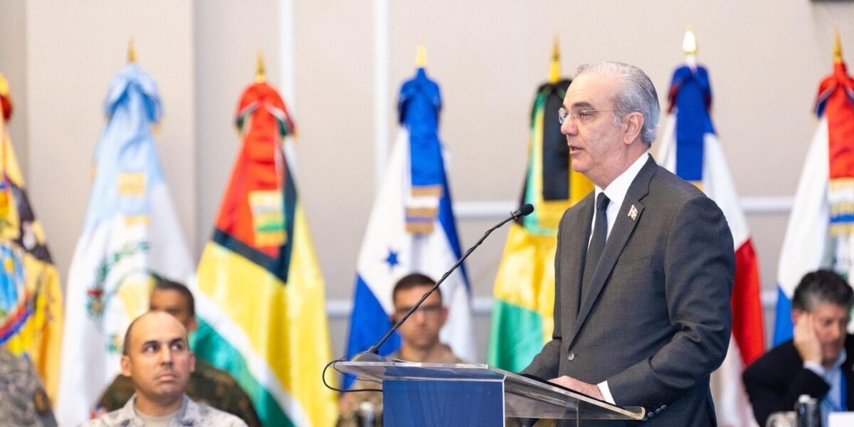 Un hombre de cabello canoso y traje gris habla en un atril transparente, delante de banderas de varios países americanos y de militares sentados.