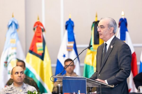 Un hombre de cabello canoso y traje gris habla en un atril transparente, delante de banderas de varios países americanos y de militares sentados.