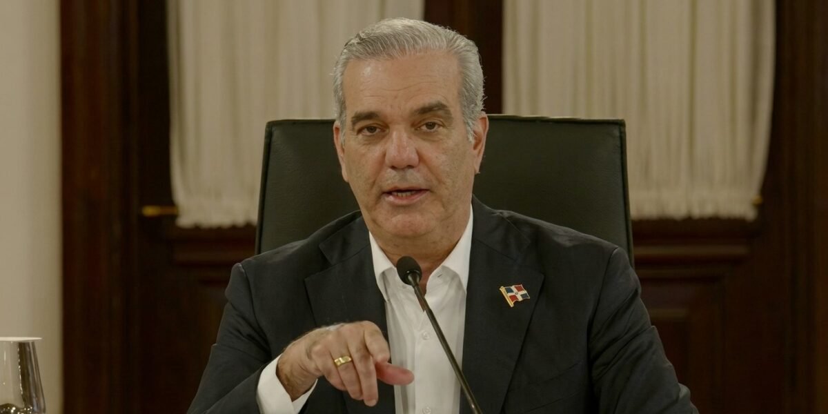 El presidente Luis Abinader habla ante un micrófono, sentado en una silla de cuero negro, con un pin de la bandera dominicana en la solapa y cortinas blancas al fondo