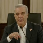 El presidente Luis Abinader habla ante un micrófono, sentado en una silla de cuero negro, con un pin de la bandera dominicana en la solapa y cortinas blancas al fondo
