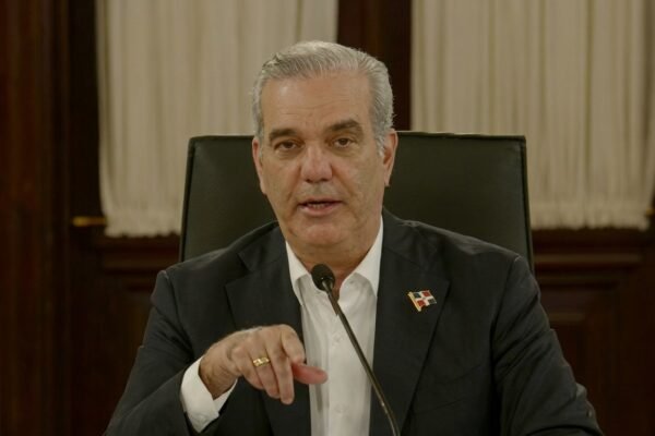 El presidente Luis Abinader habla ante un micrófono, sentado en una silla de cuero negro, con un pin de la bandera dominicana en la solapa y cortinas blancas al fondo