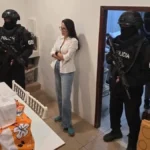 Luisa González, con camisa blanca y jeans, de pie en una vivienda junto a agentes encapuchados de la Policía de Ecuador