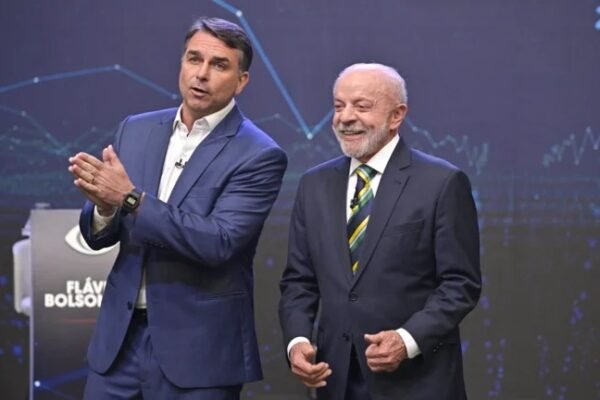 Flávio Bolsonaro, de traje azul, gesticula junto a Luiz Inácio Lula da Silva, sonriente y con corbata verde y amarilla, en un plató de televisión