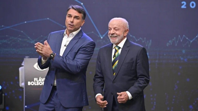Flávio Bolsonaro, de traje azul, gesticula junto a Luiz Inácio Lula da Silva, sonriente y con corbata verde y amarilla, en un plató de televisión