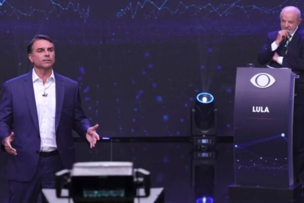 Flávio Bolsonaro habla de pie en un plató de televisión mientras Lula escucha junto a un atril con su nombre