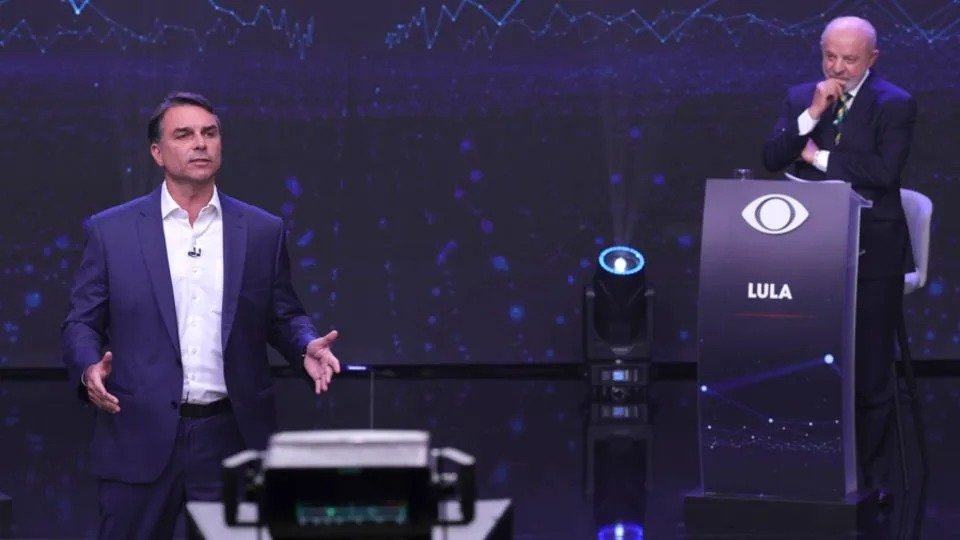 Flávio Bolsonaro habla de pie en un plató de televisión mientras Lula escucha junto a un atril con su nombre