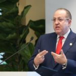 Magín Díaz habla desde un atril con micrófono, con una pantalla y una planta al fondo