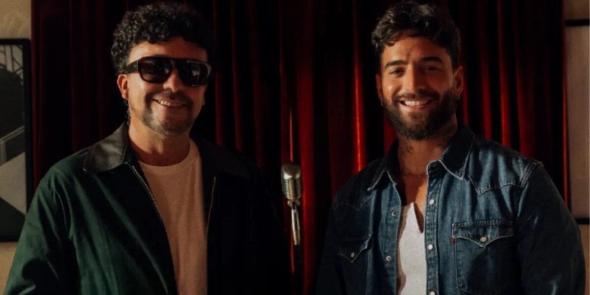 Andrés Cepeda, con gafas oscuras, y Maluma, con camisa de mezclilla, sonríen frente a un micrófono antiguo y una cortina roja