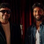 Andrés Cepeda, con gafas oscuras, y Maluma, con camisa de mezclilla, sonríen frente a un micrófono antiguo y una cortina roja