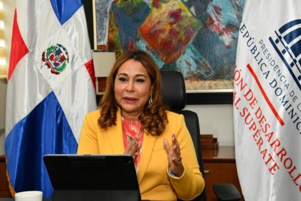 Mujer con chaqueta amarilla habla sentada ante una tableta, entre la bandera dominicana y una bandera de Supérate