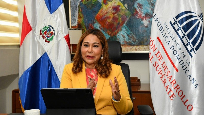 Mujer con chaqueta amarilla habla sentada ante una tableta, entre la bandera dominicana y una bandera de Supérate