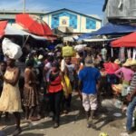 Multitud de compradores y vendedores en un mercado al aire libre, algunos con bultos en la cabeza, entre carpas y sombrillas rojas; al fondo, un edificio azul con el escudo dominicano