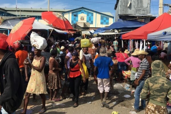 Multitud de compradores y vendedores en un mercado al aire libre, algunos con bultos en la cabeza, entre carpas y sombrillas rojas; al fondo, un edificio azul con el escudo dominicano