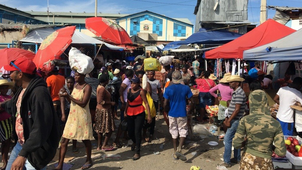 Multitud de compradores y vendedores en un mercado al aire libre, algunos con bultos en la cabeza, entre carpas y sombrillas rojas; al fondo, un edificio azul con el escudo dominicano