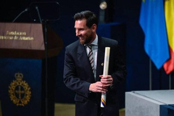 Lionel Messi, de traje oscuro, sonríe con un diploma enrollado en las manos junto al atril de la Fundación Princesa de Asturias y varias banderas.