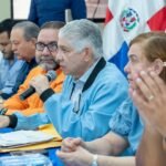 Funcionarios reunidos en una mesa con mantel azul junto a la bandera dominicana; un hombre de cabello canoso habla por micrófono