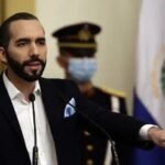 Nayib Bukele habla en un atril con micrófonos y señala con la mano; detrás, un guardia de honor con mascarilla y la bandera de El Salvador