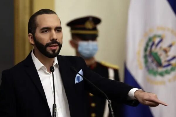 Nayib Bukele habla en un atril con micrófonos y señala con la mano; detrás, un guardia de honor con mascarilla y la bandera de El Salvador