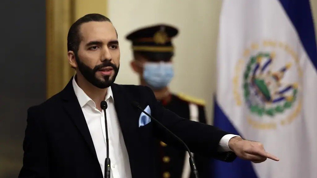 Nayib Bukele habla en un atril con micrófonos y señala con la mano; detrás, un guardia de honor con mascarilla y la bandera de El Salvador