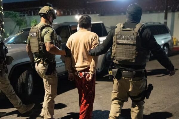Agentes federales con chalecos escoltan de noche a un hombre esposado, de espaldas, hacia un vehículo