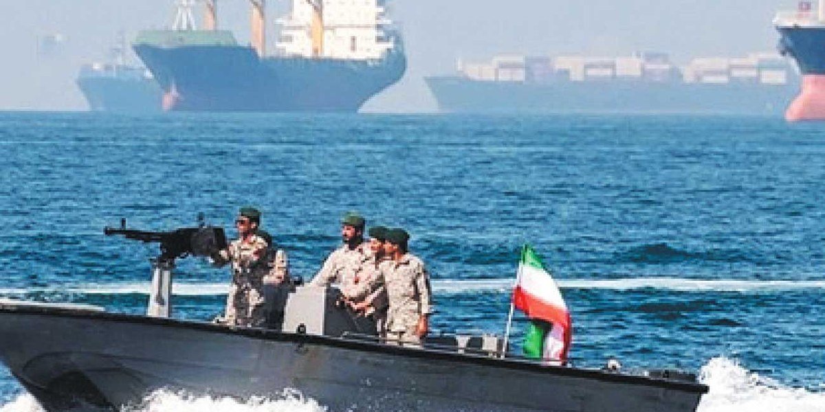 Militares iraníes en una lancha armada con bandera de Irán navegan frente a buques mercantes