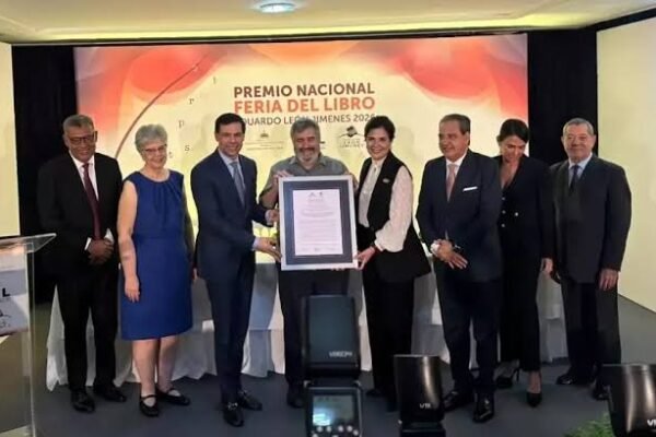 Un hombre de barba canosa con camisa gris sostiene un certificado enmarcado junto a otras personas en un escenario con el rótulo del Premio Nacional Feria del Libro Eduardo León Jimenes 2026