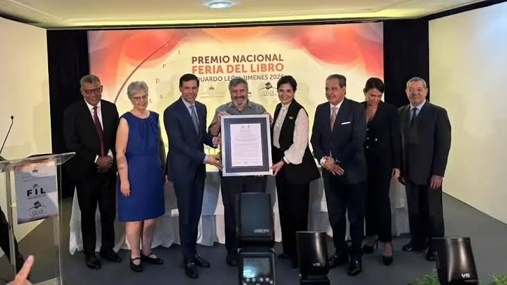 Un hombre de barba canosa con camisa gris sostiene un certificado enmarcado junto a otras personas en un escenario con el rótulo del Premio Nacional Feria del Libro Eduardo León Jimenes 2026