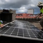 Dos técnicos con chalecos reflectantes amarillos instalan paneles solares en la azotea de un edificio, con nubes y edificios al fondo