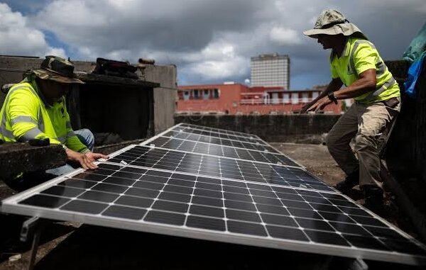 Dos técnicos con chalecos reflectantes amarillos instalan paneles solares en la azotea de un edificio, con nubes y edificios al fondo